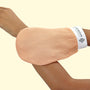 The photo shows a person wearing a Moroccan exfoliating glove on the arm. The glove has a pale pink color and is designed to cover the hand and wrist, leaving the fingers free. Printed on the white band ending around the wrist is the 'Legacy of MOROCCO' logo. The background is a uniform beige color, highlighting the contrast between the texture of the glove and the wearer's skin, suggesting delicacy and effectiveness