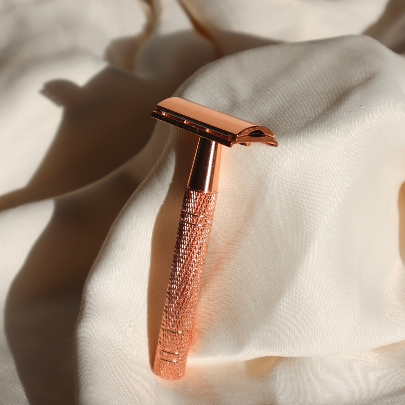 The picture shows a classic long-handled shaver set against a background of white fabric. The razor has a rose-gold metallic finish that reflects light, suggesting quality and elegance. The design is simple yet sophisticated with a detailed texture on the handle for a better grip. The razor's shadow is visible on the fabric, adding depth to the scene and accentuating its slim, stylized shape."