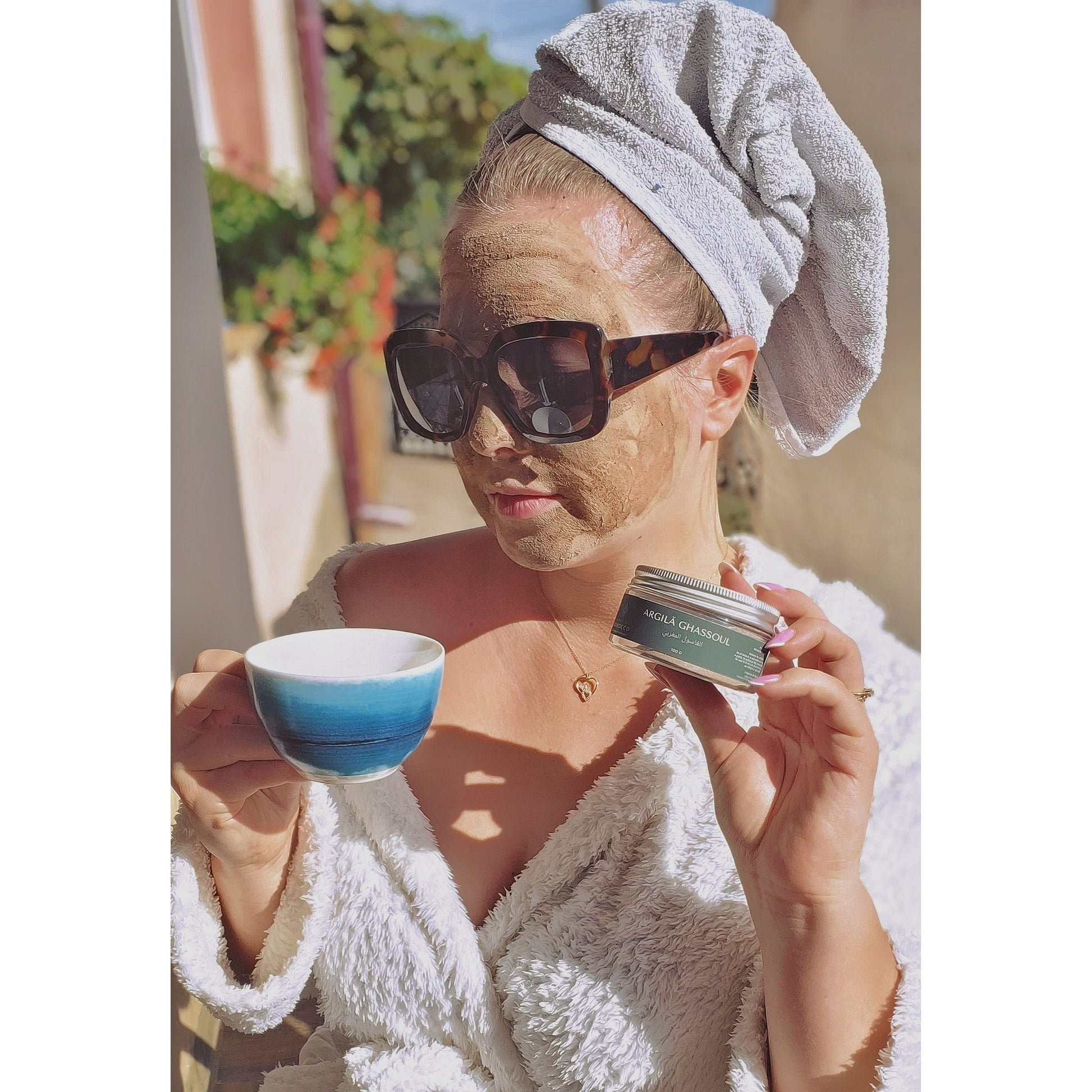 The photograph captures a woman in a white bathrobe, with a towel wrapped around her head, wearing large sunglasses, and with a clay mask applied to her face. She holds a cup of coffee or tea in one hand and a container of 'GHASSOUL CLAY' in the other. The scene is set on a bright terrace in the sunshine, suggesting a relaxing morning or an outdoor beauty ritual.