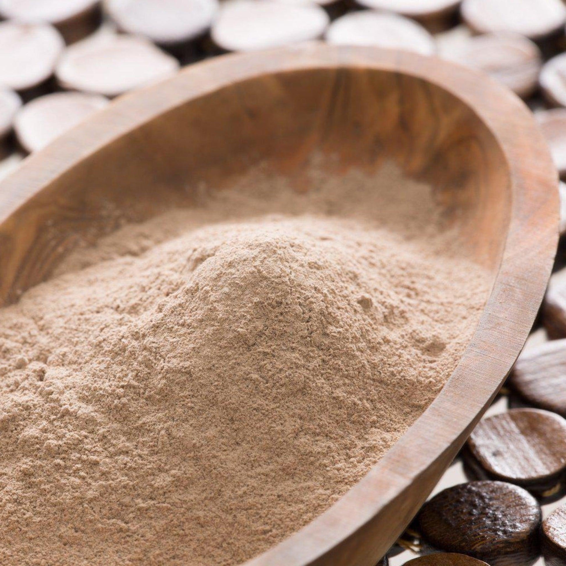 The image shows a generous amount of finely ground, beige-colored powder placed in an oval wooden bowl, with the natural texture of the wood visible. The bowl is set against a dark-toned wooden background and the powder has a smooth, homogenous texture, suggesting a natural product, possibly for use in skin care rituals or for culinary purposes.